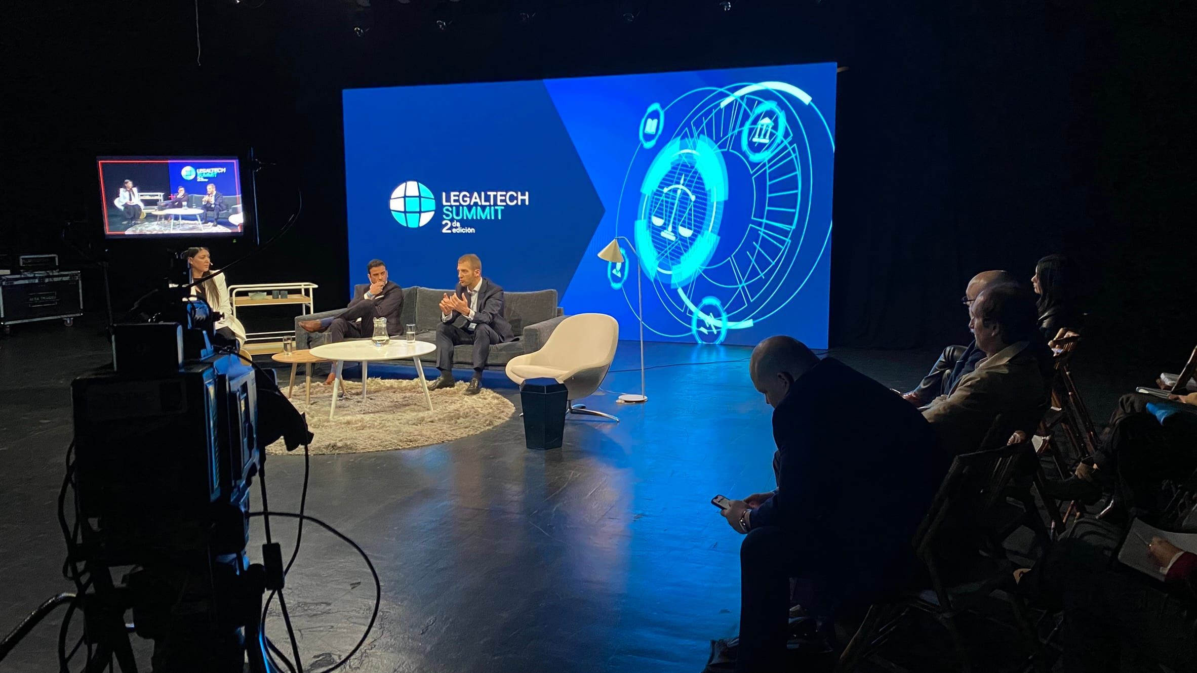 Set de estudio durante el Legaltech Summit: panel de dos invitados frente a una pantalla LED, con una cámara de estudio en primer plano y público sentado